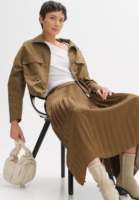 Veste marron courte avec deux poches avant, débardeur blanc et jupe maxi plissée marron. Tient un sac de couleur crème. Bottines beiges.