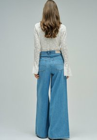 Mujer con cabello largo y castaño, vistiendo una blusa blanca de manga larga con encaje y pantalones vaqueros azules de pierna ancha y talle alto, de pie contra un fondo liso.