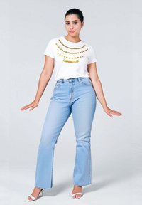 T-shirt bianco con un motivo di perline dorate, abbinato a jeans a zampa leggeri di colore blu chiaro. Il modello indossa sandali bianchi a punta aperta. Sfondo semplice.