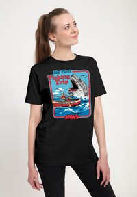 Mladá žena v černém tričku s kresleným motivem dvou lidí rybařících na loďce a velkým žralokem vyplouvajícím z vody za nimi.