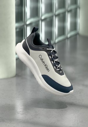 Lichtgrijze en donkerblauwe Calvin Klein sneaker met witte zool en veters, zwevend boven een gepolijste betonnen vloer, glazen wand op de achtergrond.