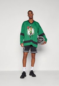 Zöld hosszú ujjú mez fekete részletekkel, Boston Celtics grafikával. Fekete rövidnadrággal, fehér zoknival és fekete lábbelivel párosítva.
