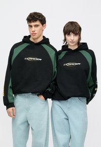 Mustad kapuutsid roheliste detailidega, millel on kiri "yTRACER." Sobitatud helesiniste lahtiste teksadega. Lihtne, vabaaja disain.