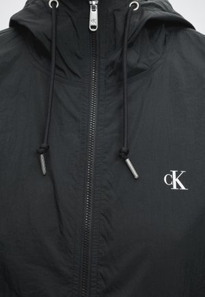 Chaqueta negra con capucha con cremallera frontal, cordones negros con puntas metálicas y logo blanco "cK" en el lado derecho del pecho.