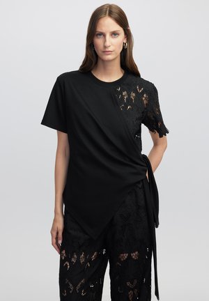 Camiseta negra con un panel de encaje que presenta estampados florales, mangas cortas y un diseño cruzado, combinada con pantalones de encaje negros.