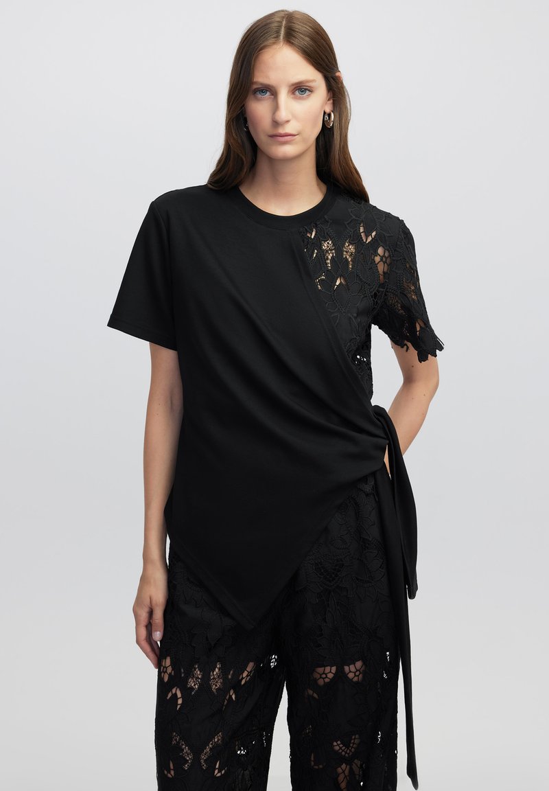 Camiseta negra con un panel de encaje que presenta estampados florales, mangas cortas y un diseño cruzado, combinada con pantalones de encaje negros.