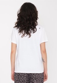 Hvid bomulds t-shirt med korte ærmer, afslappet pasform og et lille logo på den øverste ryg. Båret med mønstrede sorte shorts.