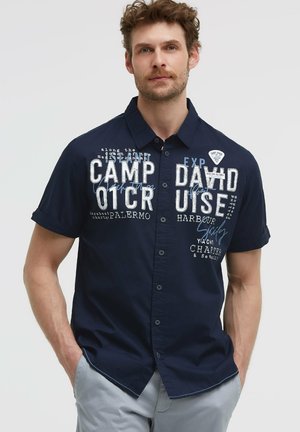 Mann mit lockigem Haar, der ein marineblaues kurzärmliges Hemd mit weißem und blauem grafischem Text und hellgraue Hose trägt.