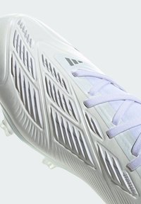 Scarpa da calcio bianca con materiale testurizzato, forma aerodinamica e design simile a delle aperture. Presenta lacci viola chiaro e tacchetti trasparenti.