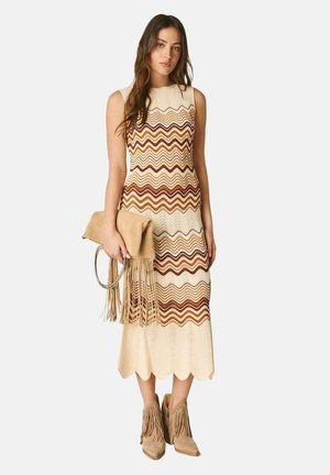 Donna che indossa un abito midi senza maniche a righe ondulate in tonalità beige e marrone, tiene una pochette cuoio con frange e indossa stivaletti con frange.