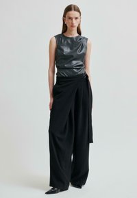 Žena v bezramenném tmavošedém koženém topu, černých širokých kalhotách s vysokým pasem a obvazovým páskem, a špičatých černých botách, stojí proti šedému pozadí.