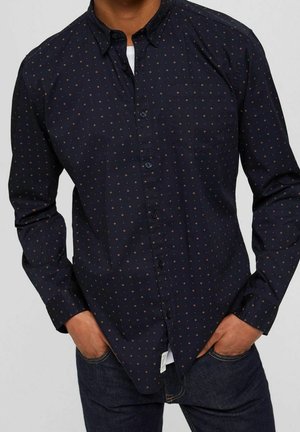 Man wearing a dark navy button-up shirt with small brown dot patterns, hands in pockets, paired with dark denim jeans.