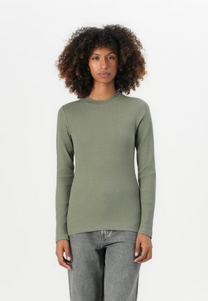 Langermet ribbestrikket topp i olivengrønn, med rund halsuka og tettsittende silhuett, kombinert med lysegrå jeans.