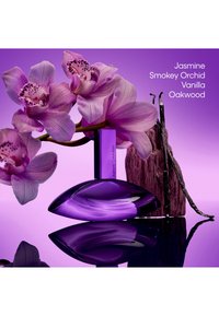 Paarse orchideeën naast een Calvin Klein Euphoria parfumflesje met vanillestokjes en hout op een paarse reflecterende ondergrond.