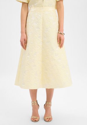 Frau trägt ein blassgelbes Midikleid aus Jacquard mit Blumenmuster, kurzen Ärmeln, einem goldenen Armband und goldenen High-Heel-Sandalen mit Knöchelriemen.