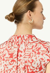 Abito a fantasia floreale nei toni del corallo e bianco, con un design a pieghe sul retro. La modella indossa orecchini a cerchio dorati e ha i capelli raccolti in un bun.
