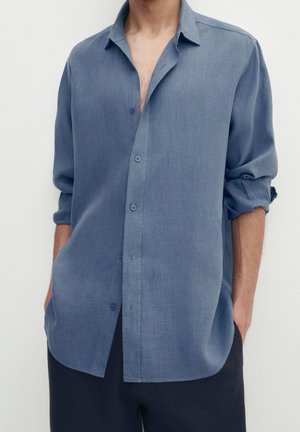 Chemise bleu à boutons en tissu léger, avec une coupe décontractée, des manches retroussées et un ourlet légèrement arrondi.
