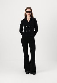 Blazer noir en tricot à double boutonnage avec des boutons métalliques, associé à un pantalon noir ajusté évasé. Le mannequin porte également des lunettes de soleil rectangulaires noires.