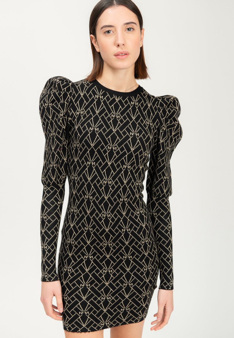 Donna che indossa un vestito nero aderente con motivi geometrici beige e spalle a palloncino, in piedi contro uno sfondo chiaro e semplice.