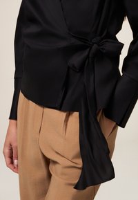 Zwarte wikkelblouse gemaakt van gladde stof, met een strik in de taille. Gepaarde met lichtbeige, gestructureerde, loszittende broek.