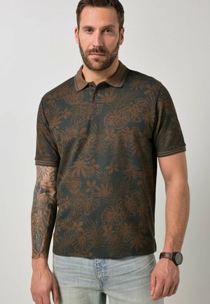 Mężczyzna z brodą, ubrany w ciemnozieloną i brązową koszulkę polo w kwiatowy wzór, jasne dżinsy, z tatuażem na lewej ręce oraz czarny zegarek na rękę z brązowym paskiem.