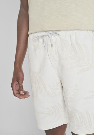 Close-up van een persoon die beige gestructureerde shorts draagt met een trekkoord en een subtiel bladmotief, en een licht beige top.