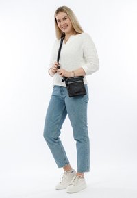 Schwarze Leder-Crossbody-Tasche mit strukturierten Oberflächen, verstellbarem Tragegurt und Reißverschlussfächern. Das Modell trägt einen weißen Pullover und blue Jeans.