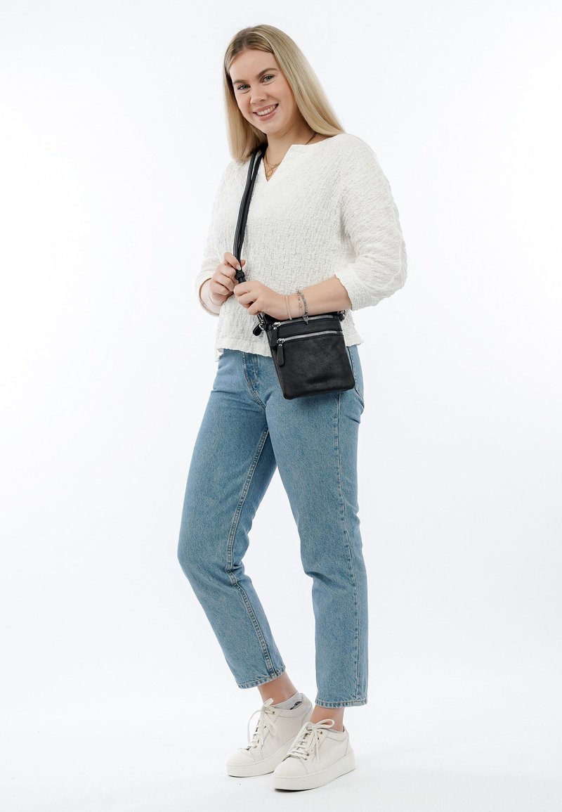 Schwarze Leder-Crossbody-Tasche mit strukturierten Oberflächen, verstellbarem Tragegurt und Reißverschlussfächern. Das Modell trägt einen weißen Pullover und blue Jeans.