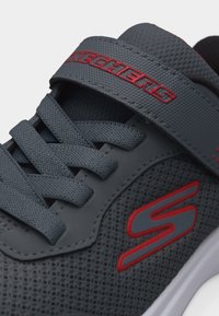 Κοντινό πλάνο σκούρου γκρι αθλητικού παπουτσιού Skechers με κόκκινο λογότυπο, υφασμάτινο δίχτυ με υφή, κορδόνια στο ίδιο χρώμα και λουράκι στο επάνω μέρος.