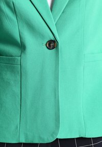 Blazer vert en tissu texturé. Dispose d'un bouton marron unique, deux poches latérales et des revers structurés.