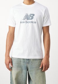 Bílé bavlněné tričko s šedým logem "NB" a textem "new balance". Krátké rukávy a klasický design s kulatým výstřihem.