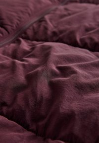 Bordeaux geknoopte stof met een zachte, licht glanzende textuur, met een zichtbare rits langs één rand en gekreukelde oppervlaktedetails.