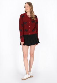 Röd stickad cardigan med svarta båg mönster, rund hals, framknäppning och långa ärmar, kombinerad med en svart plisserad kjol och vita sneakers.