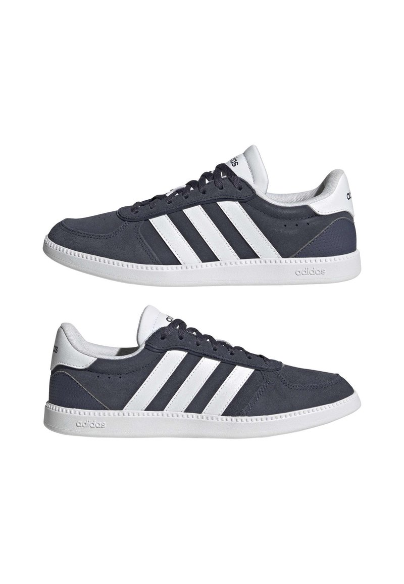 Zapatilla gris oscuro con diseño de tres rayas blancas, parte superior de ante, suela de goma blanca y forro de tela. Presenta un cierre de cordones.