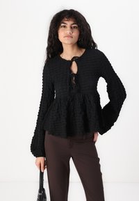 Black peplum top with textured fabric, long bell sleeves, and a front tie closure. Worn with brown trousers and holding a black bag.