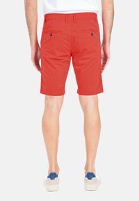 Pantaloni corti rossi in cotone con una texture liscia, dotati di due tasche posteriori e di accenti in blu a contrasto sui bottoni. Finiti all'altezza della metà della coscia.