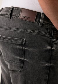 Jeans de mezclilla negros para hombre con una etiqueta de cuero "MEN+", bolsillo trasero y textura de tela suave con un ligero desgaste y detalles en la costura.