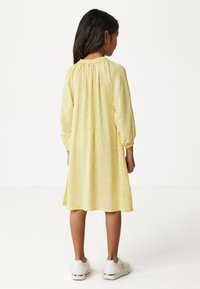 Gelbes Blumenkleid mit langen Ärmeln, gerafftem Ausschnitt und fließender Silhouette. Getragen mit weißen Sneakern mit dekorativen Akzenten.