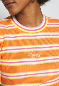 Pomarańczowa bluzka z prążkowanej tkaniny z poziomymi paskami w kolorze różowym, żółtym i białym. Białe logo "Tommy Jeans" na klatce piersiowej, z kolorowym prążkowanym dekoltem.