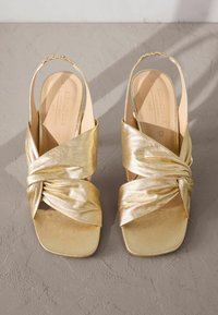 Gouden metallic hoge hakken sandalen met een geknoopte ontwerp, open teen en slingback band. Gladde textuur en vierkante vorm.