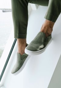 Zapatillas de ante verde sin cordones con una suela de goma blanca, que presentan detalles de costura y una textura suave. Mostradas sobre un fondo claro.