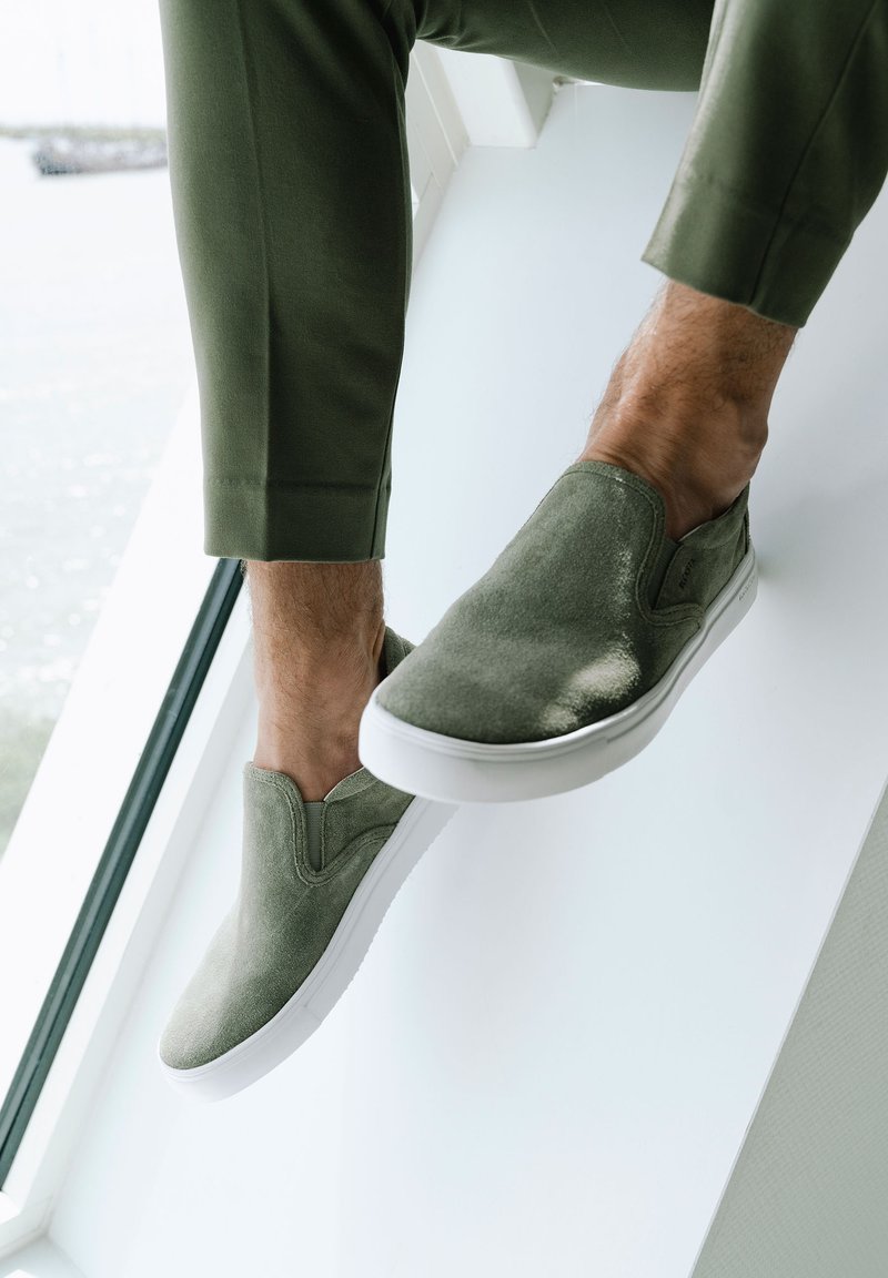 Zapatillas de ante verde sin cordones con una suela de goma blanca, que presentan detalles de costura y una textura suave. Mostradas sobre un fondo claro.