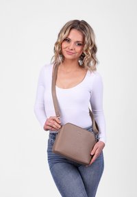 Žena s vlnitými blond vlasy nosí bílé tričko s dlouhým rukávem a modré džíny, drží taupe kabelku přes rameno s logem na přední straně.