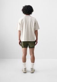 Beige bomulls t-shirt med korta ärmar, parat med olivgröna shorts. Modellen har på sig vita strumpor och beige sneakers, sedd bakifrån.