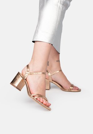 Sandalias de tacón en metalizado rosa dorado con doble tira y tacón cuadrado. Textura suave y detalle de tira ajustable en el tobillo.