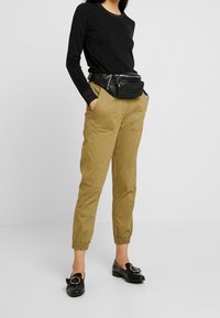 Zwarte lange mouwen top, tan cargobroek met elastische boorden en zwarte platte schoenen met een ronde accent. Een zwarte heuptas wordt om de taille gedragen.