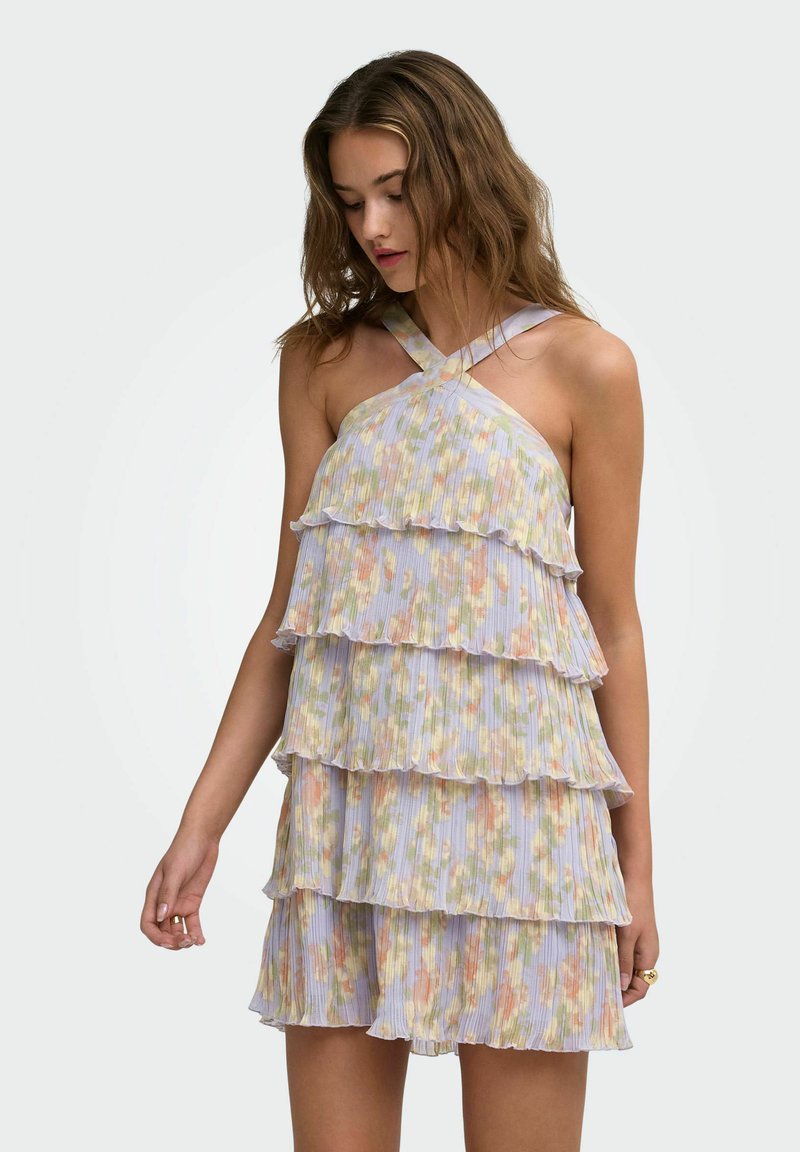 Jeune femme portant une robe sans manches à volants florale avec des bretelles croisées, regardant vers le bas, debout devant un fond uni.