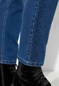 Vaqueros de mezclilla azul con un ajuste slim, con una fila de cuentas decorativas negras a lo largo de la costura lateral. Combinados con botas negras de terciopelo con cordones.