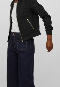 Veste bomber noire avec des zips dorés, portée sur un débardeur blanc. Associée à un jean en denim bleu foncé, ample, avec des coutures orange.