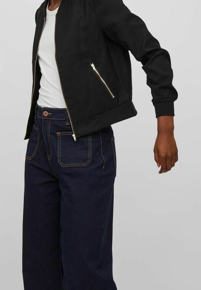 Veste bomber noire avec des zips dorés, portée sur un débardeur blanc. Associée à un jean en denim bleu foncé, ample, avec des coutures orange.